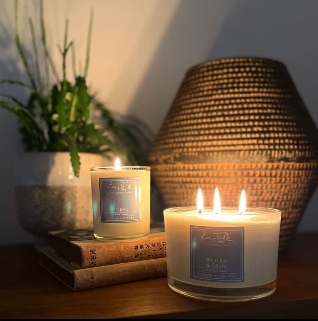 Eve Taylor London aromatherapy candles glowing on a dark wood surface beside books, a plant, and soft lamp light.