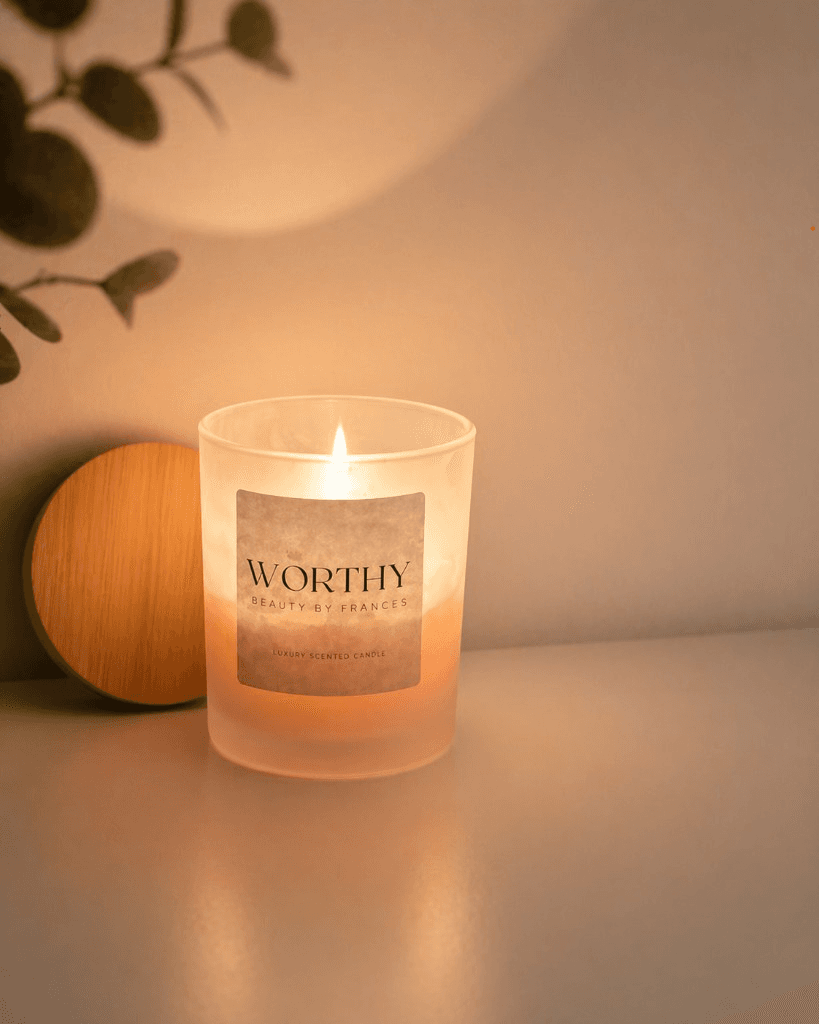 Worthy Beauty luxury scented candle in frosted glass with a warm flame, wooden lid, and soft eucalyptus in a bright, minimal setting.