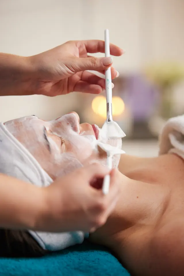 Close-up of a guest receiving a facial while a practitioner applies a white mask with soft brushes in a warm, candlelit room.
